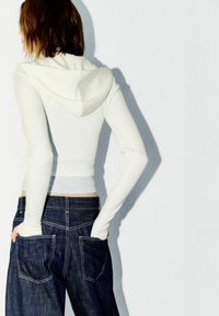 Cream hooded sweatshirt with long sleeves, cropped design, paired with dark blue denim jeans featuring back pockets and contrast stitching.