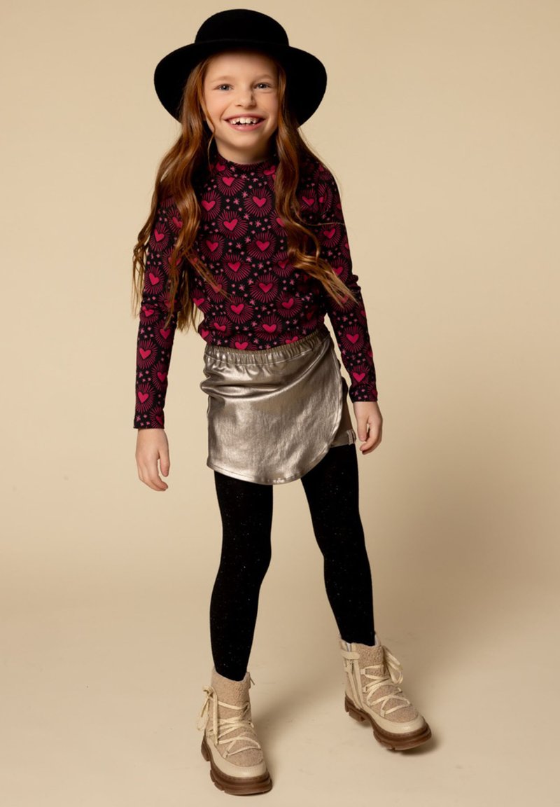 Glimlachend meisje met een zwarte hoed, een zwart longsleeve shirt met roze harten, een metallic zilveren rok, zwarte leggings en beige winterlaarzen.