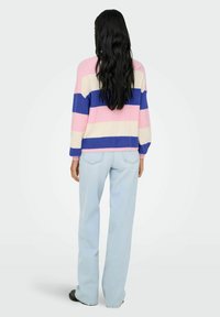 Femme aux longs cheveux noirs portant un pull rayé pastel et un jean bleu clair, debout de dos devant un fond clair uni.