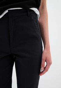 La main droite reposant le long du corps d'une personne portant un pantalon noir avec une poche zippée et des hauts noirs et blancs superposés sur un fond uni.