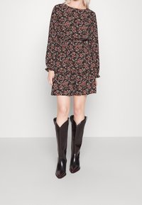 Vestido negro con flores, de manga larga y dobladillo con volantes, combinado con botas de cowboy de cuero marrón hasta la rodilla, con punta afilada y parte superior en forma de V.