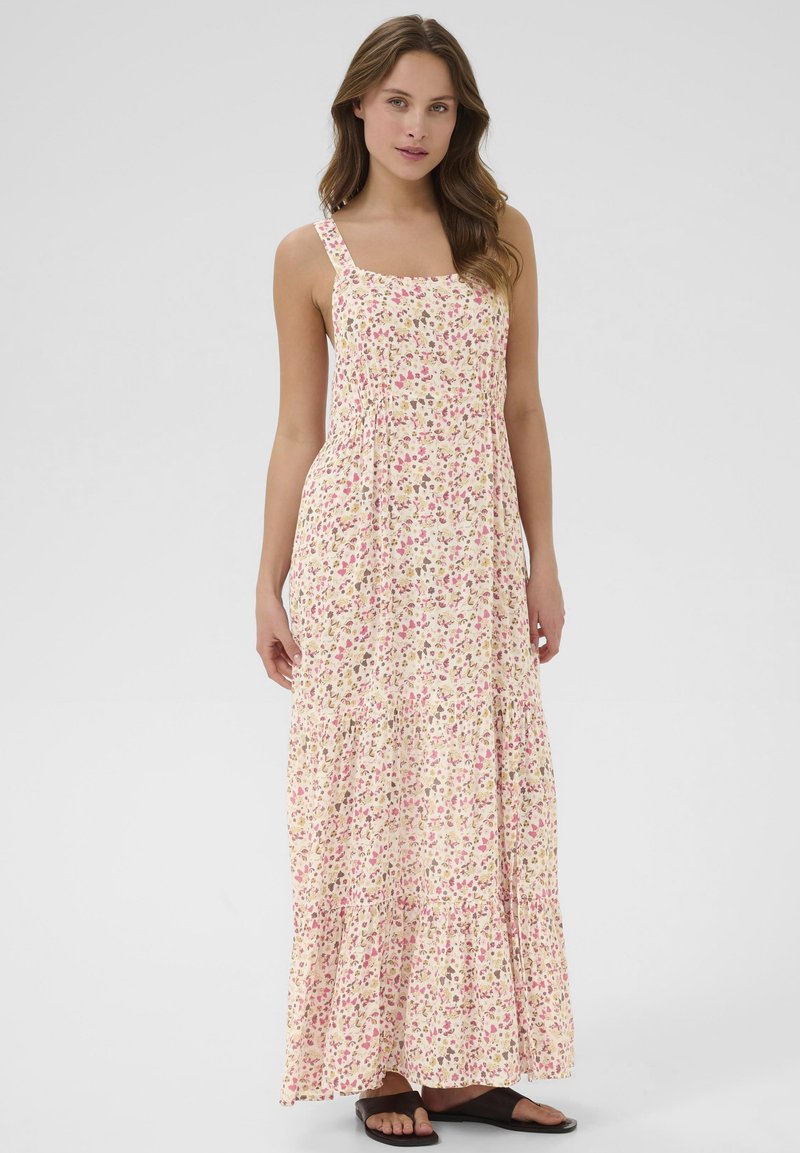 Kvinde iført ærmeløs, ankellang blomstret kjole i bløde rosa, beige og brune nuancer med sorte flade sandaler, stående mod en ensfarvet baggrund.