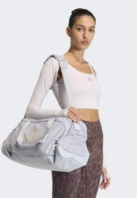 Ανοιχτό γκρι σακ βουαγιάζ με αξεσουάρ από δίχτυ, διπλές λαβές και ρυθμιζόμενο ιμάντα. Διαθέτει τρεις διαγώνιες ρίγες και διάτρητες λεπτομέρειες.