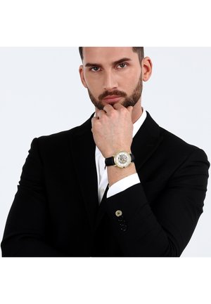 Gold and silver watch with black leather strap, round face displaying intricate details. Model's hand rests on chin while wearing a black suit jacket.