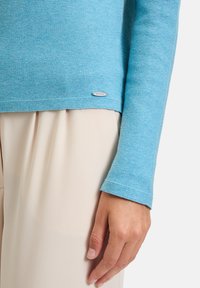 Hellblauer Pullover aus strukturiertem Stoff. Mit langen Ärmeln und einem kleinen metallenen Etikett mit "BETTAG." Kombiniert mit einem cremefarbenen Unterteil.