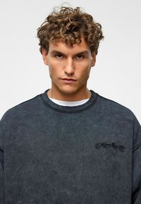 Zwarte sweatshirt met een gestructureerde afwerking, ribbelkraag en geborduurd detail op de borst. Relaxte pasvorm en lange mouwen.