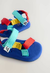 Blå sandaler med färgglada remmar i röd, gul och akvamarin; tillverkade av mjukt material med en strukturerad sula för bättre grepp. Lättviktsdesign.