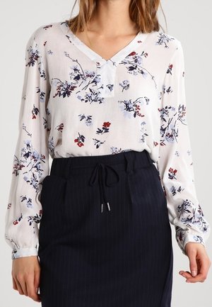 Femme portant un chemisier à manches longues blanc à motif floral avec un col en V, rentré dans une jupe taille haute noire à fines rayures avec une taille à cordon.
