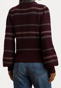 Kobieta w bordowym swetrze dzianym w białe zygzaki i niebieskich dżinsach, pokazana z tyłu.