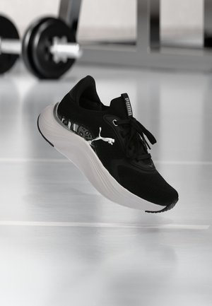 Baskets Puma noires avec semelle blanche et logo, flottant au-dessus d'un sol de gym avec des poids flous en arrière-plan.