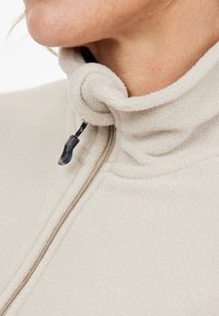 Primo piano del colletto di un giaccone in pile beige con tirazip nero e cordino regolabile vicino al collo di una persona.