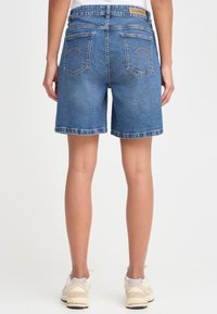 Vidutinio atspalvio džinsiniai šortai su tiesiu kirpimu, suveltu kraštu ir tradicinėmis galinėmis kišenėmis. Ant juosmens matomas etiketės ženklelis.