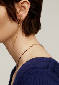 Pendiente de oro con un detalle de cuentas coloridas, con una cadena que cae hacia abajo. Collar corto de cuentas multicolores, suéter de textura azul oscuro.