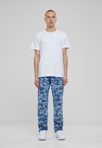 Camiseta blanca de manga corta combinada con pantalones de camuflaje azul y zapatillas blancas, de pie contra un fondo neutro.