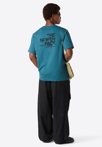 Hombre vestido con una camiseta teal de The North Face, pantalones anchos negros, gafas de sol y llevando un pequeño bolso cruzado beige, de pie de espaldas.