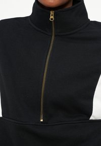 Κοντινό πλάνο μαύρου ρούχου με ψηλό γιακά και χρυσό φερμουάρ που κλείνει μερικώς, εμφανίζοντας ένα μικρό τμήμα λευκού υφάσματος στο πλάι.