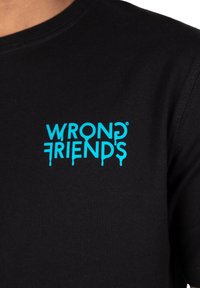 Camiseta de algodón negra con un logo turquesa "WRONG FRIENDS" con efecto de goteo, situado en la parte superior izquierda del pecho.