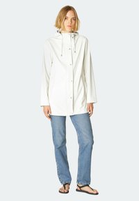 Chaqueta blanca impermeable con capucha, cierre de botones en la parte delantera, dos bolsillos laterales y cordones; llevada sobre unos vaqueros azules y sandalias negras.