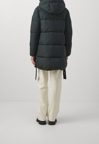 Black puffer coat with a hood, quilted design, adjustable drawstrings at the hem, worn over cream-colored pants. Textured, insulated fabric.