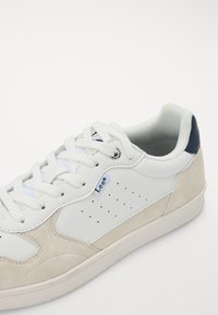 Zapatilla de cuero blanca con acentos de ante beige, puntera redonda, suela plana y detalle perforado. Presenta cordones blancos y un acento en el talón azul.