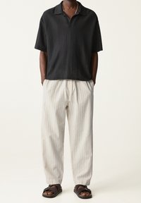 Camiseta polo negra de manga corta con tejido texturizado, combinada con pantalones relajados de rayas crema y sandalias oscuras.