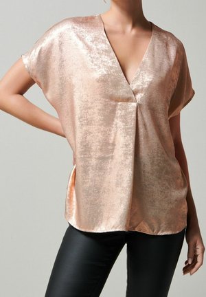 Blouse manches courtes en or rose, tissu brillant et texturé, col en V et coupe ample. Associée à un pantalon noir ajusté.