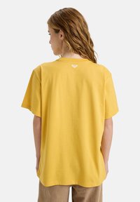 Geel katoenen t-shirt met een losse pasvorm, korte mouwen en een ronde halslijn. Heeft een klein logo op de achterkant van de hals.