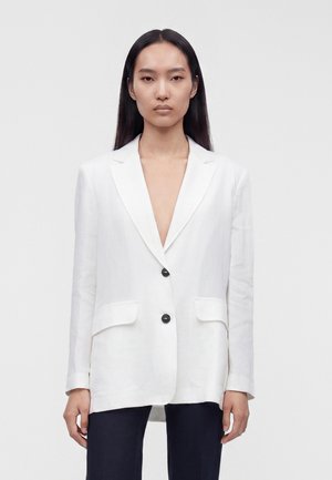 Vrouw met steil zwart haar draagt een witte blazer met twee zwarte knopen en een donkere broek, staand tegen een effen lichte achtergrond.