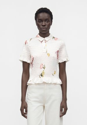 Vrouw met kort krullend haar draagt een witte gesmokte blouse met korte mouwen, bloemenprint en een gerimpelde zoom, gecombineerd met witte broek, staand tegen een effen achtergrond.