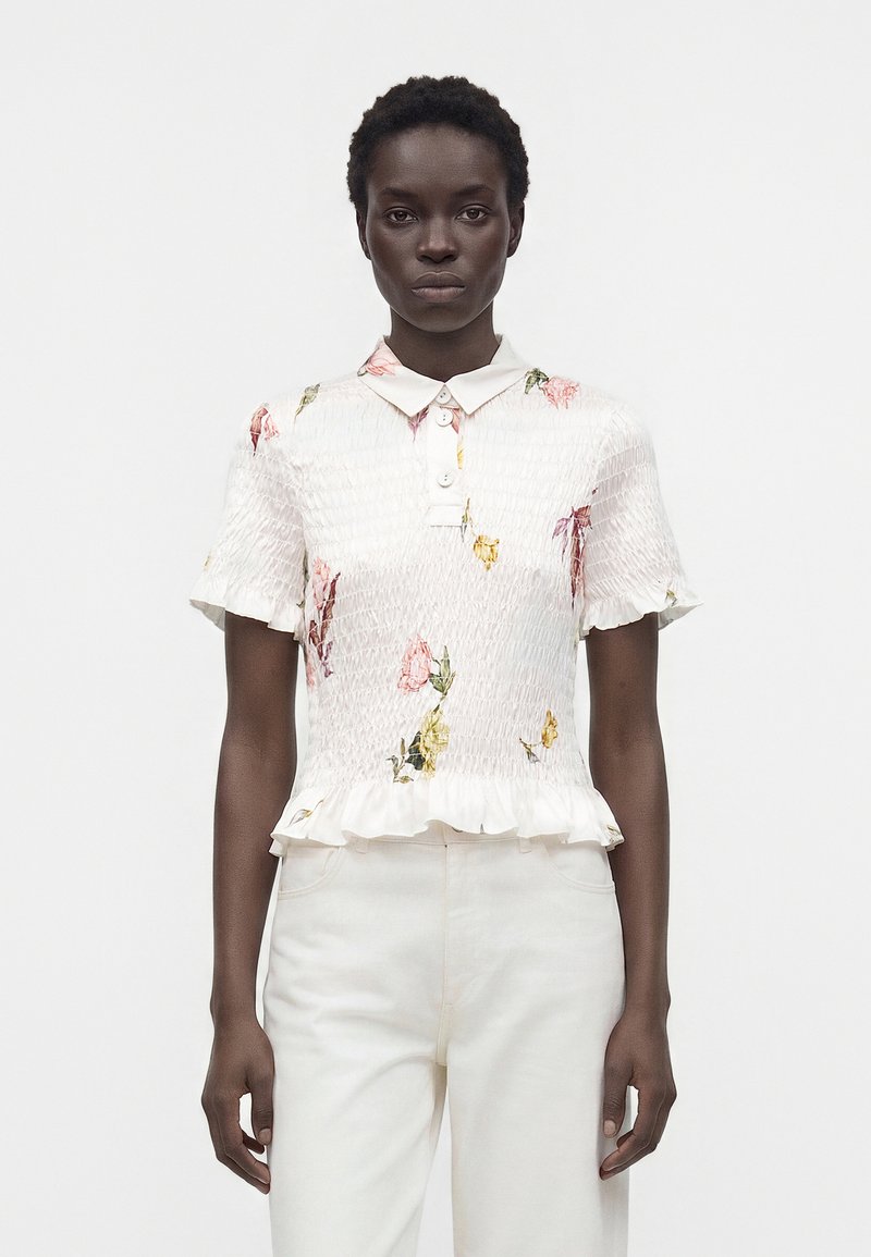 Femme aux cheveux courts et bouclés portant un chemisier blanc smocké à manches courtes avec un imprimé floral et un ourlet à volants, associé à un pantalon blanc, debout devant un fond uni.