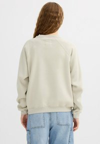 Sudadera de color gris claro con un corte holgado, mangas raglán y puños acanalados. Presenta un pequeño parche cuadrado en la parte trasera del cuello. Diseño casual.