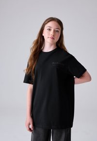 Hosszú hajú fiatal, fekete AllSaints pól�ót és sötét nadrágot visel, egyszínű világosszürke háttér előtt áll.