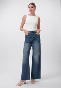 Hvit utenermet topp kombinert med høyopptrukne, vide blå jeans med falmet vask og frynsete kanter, iført strappy hvite sandaler.