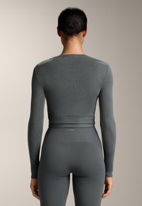 Dugi rukav, prilagođeni top i tajice visokog struka od tamnosivog rebrastog materijala. Uključuje detalj struka s naborom i logo brenda na pojasu.