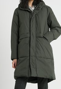 Abrigo acolchado de color verde oliva, hasta la rodilla, con capucha, cierre frontal de botones y cintura ceñida por un cinturón. Tejido texturizado con secciones acolchadas e insulated.