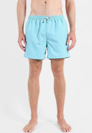 Homme portant un short de bain bleu clair avec un cordon blanc, debout pieds nus devant un fond clair uni.