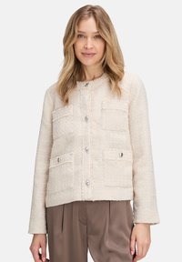 Veste beige texturée avec quatre poches à l'avant, boutons en métal argenté et col rond. Manches longues avec une coupe droite. Tissu doux et tissé.