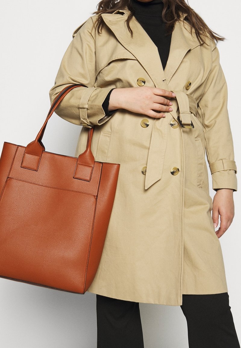 Vrouw in beige trenchcoat die een grote bruine lederen tas vasthoudt, gekleed in een zwarte broek en een zwart bovenstuk, tegen een witte achtergrond.
