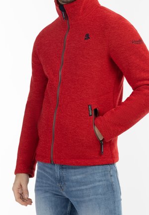 Homme portant une veste polaire rouge à fermeture éclair avec la main dans la poche zippée et un jean bleu, montré du cou aux hanches sur fond blanc.