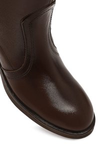 Botín de cuero marrón con acabado brillante, puntera redondeada, detalles de costura en contraste y sutilezas de costura lateral para un diseño clásico.