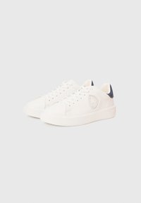 Ζευγάρι λευκά χαμηλά αθλητικά παπούτσια με κορδόνια, υφασμένη σόλα και μαύρη ετικέτα στη φτέρνα σε απλό ανοιχτό φόντο.