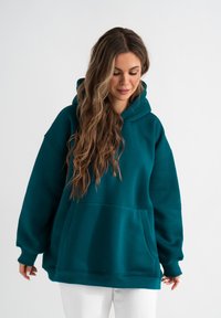 Felpa over-size color verde acqua, in misto cotone, con una grande tasca frontale, cappuccio con coulisse e polsini a costine, indossata con pantaloni bianchi.