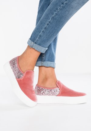 Person trägt hochgekrempelte blaue Jeans und pinke Slip-On-Sneakers mit glitzernden Fersenpartien und dicken weißen Sohlen auf weißem Hintergrund.