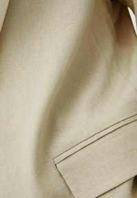 Beżowa tkanina lniana o gładkiej fakturze, z detalem w postaci złożonej kieszeni. Powierzchnia ma subtelny wzór tkania.