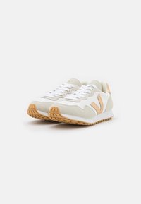 Hvite sneakers med lys grå og ferskenfarge detaljer, mesh- og semsket skinnmaterialer, flate lisser og en teksturert gummisåle.