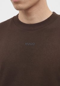Мъж, носещ кафява риза с кръгло деколте, с отпечатано малко име на марката HUGO на гърдите.