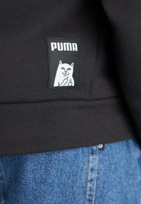 Sort sweatshirt med et PUMA-logo patch, der har et hvidt kattebillede. Ribbet detalje ved bunden. Blå denim synlig nedenunder.