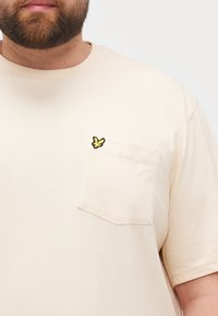 Beige bomull t-skjorte med korte ermer og en brystlomme som har en gul brodert sommerfugl-logo. Glatt tekstur.