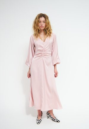 Donna con capelli mossi che indossa un lungo vestito rosa pallido con vita arricciata e maniche lunghe, abbinato a décolleté basse con stampa zebra bianco e nero.