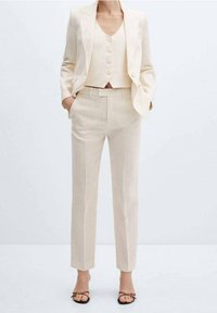 Costume en lin beige comprenant un blazer ajusté avec des revers crantés, un gilet court à boutons et un pantalon fuselé. Texture lisse, design minimaliste.